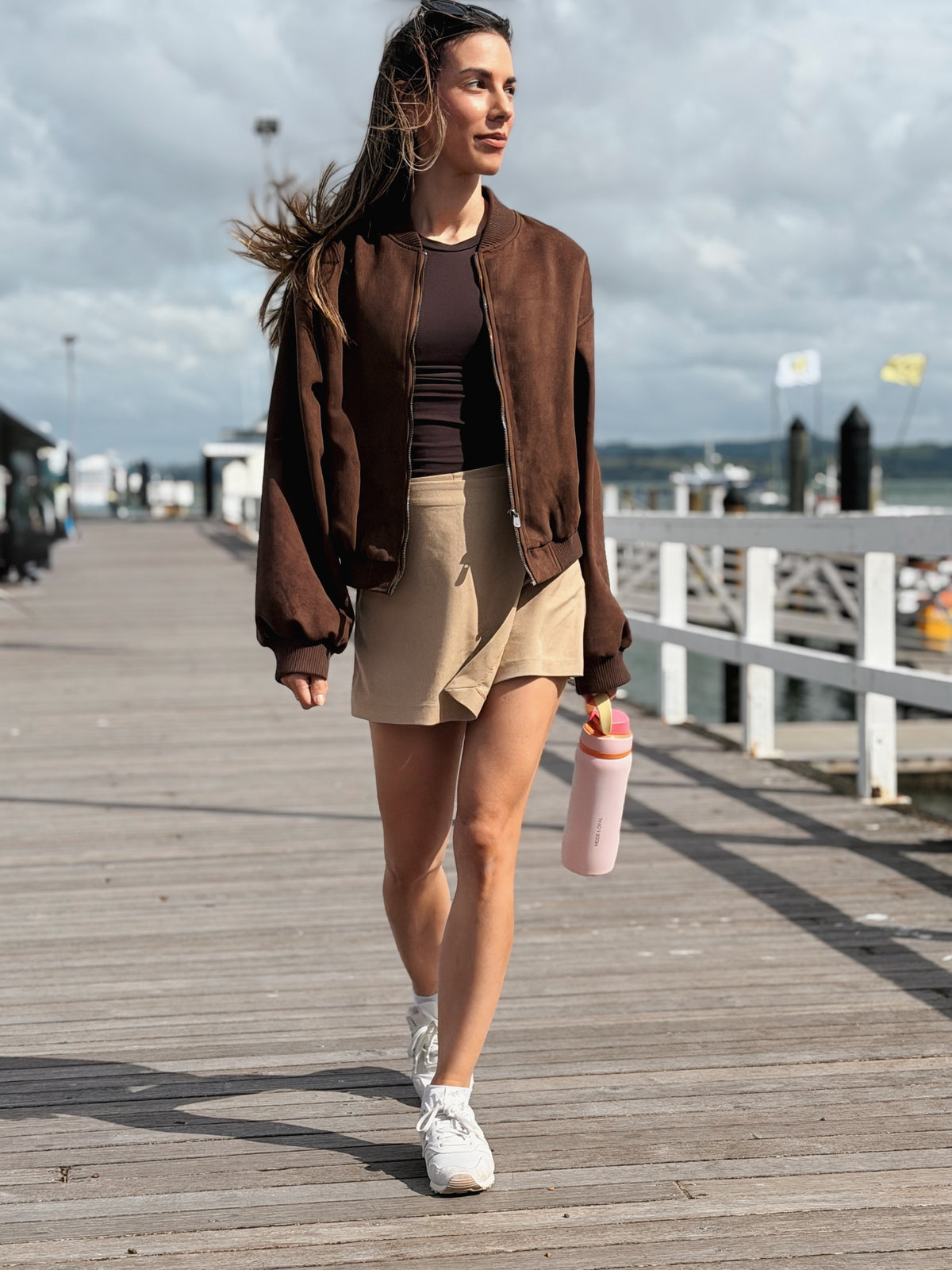 Woman in a brown blazer walking along a boardwalk with a Petal Pink Mode & Opal bottle.